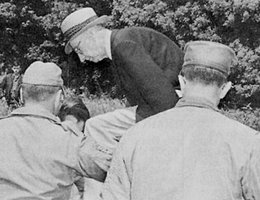 Internationally known peace activist A. J. Muste (in brimmed hat at left) initiates civil disobedience at Mead Missile Base by climbing over a gate after having been refused permission to enter; Ross Anderson (right) awaits his turn